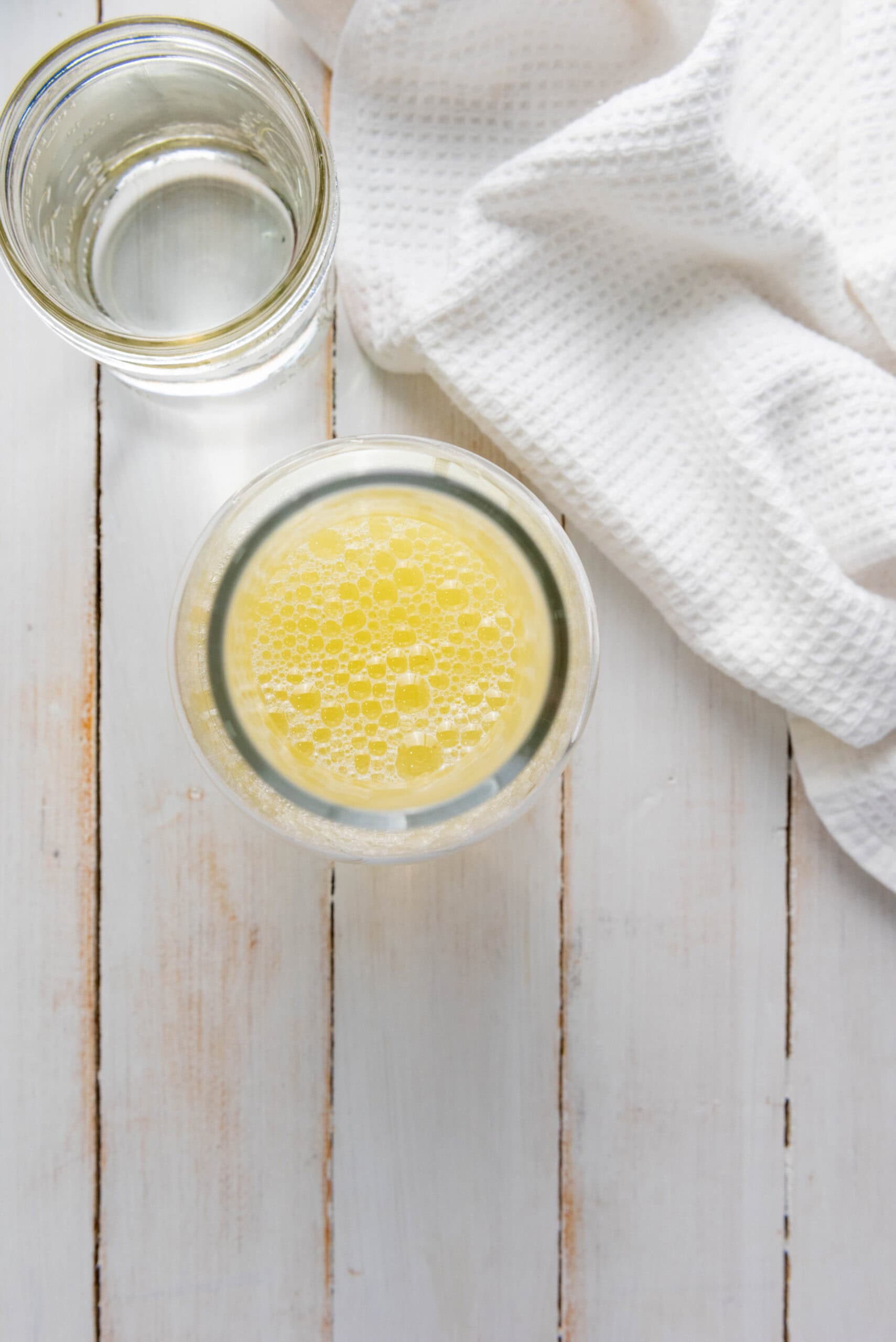 A glass of yellow homemade lemonade and a glass of water on a white wooden table with a textured white cloth nearby.