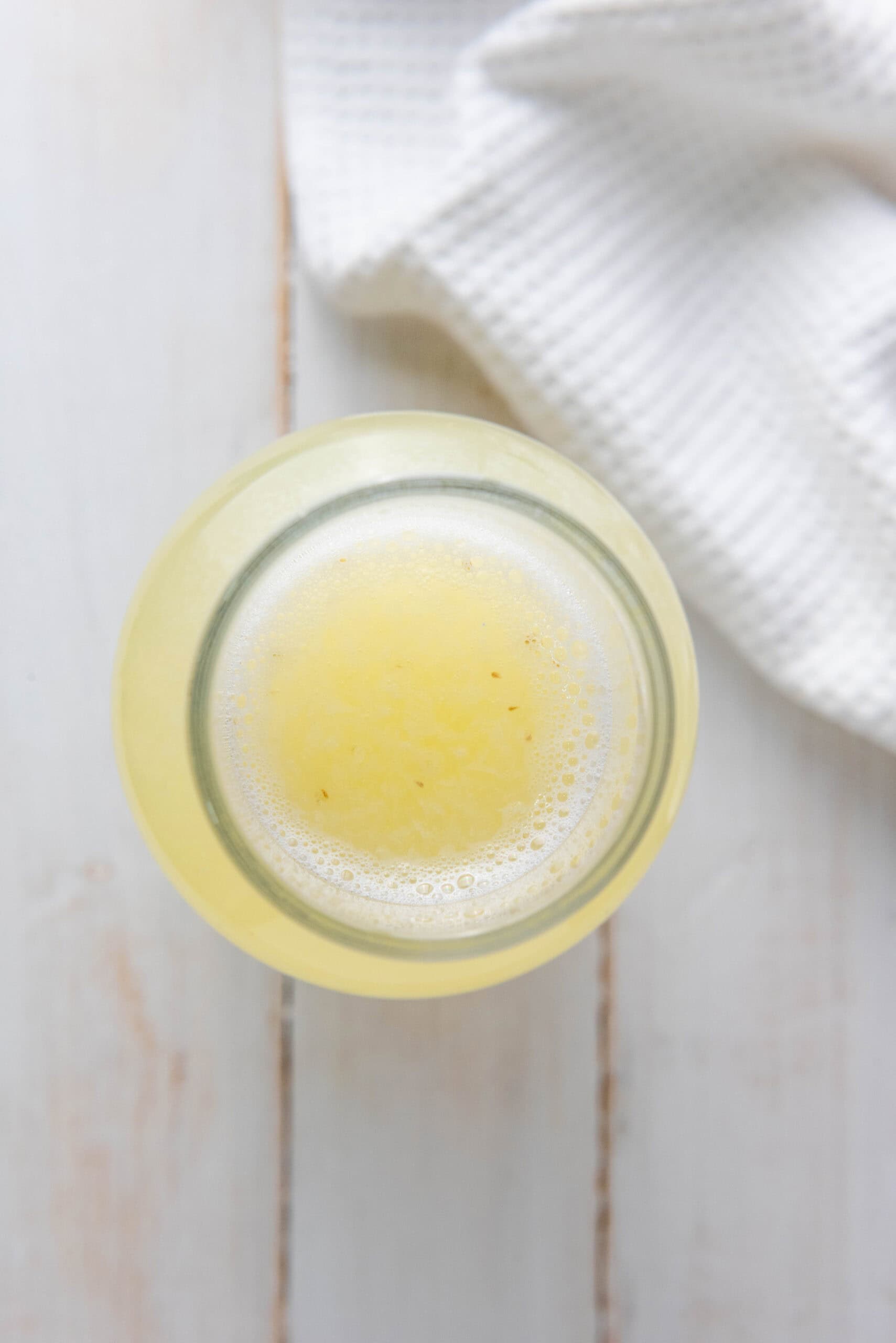 A glass of light yellow homemade lemonade sits on a white wooden table with a white textured cloth nearby.