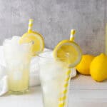 Two glasses of iced lemonade with lemon slices and yellow striped straws are pictured, alongside whole lemons in the background—perfect for enjoying a refreshing homemade lemonade recipe.