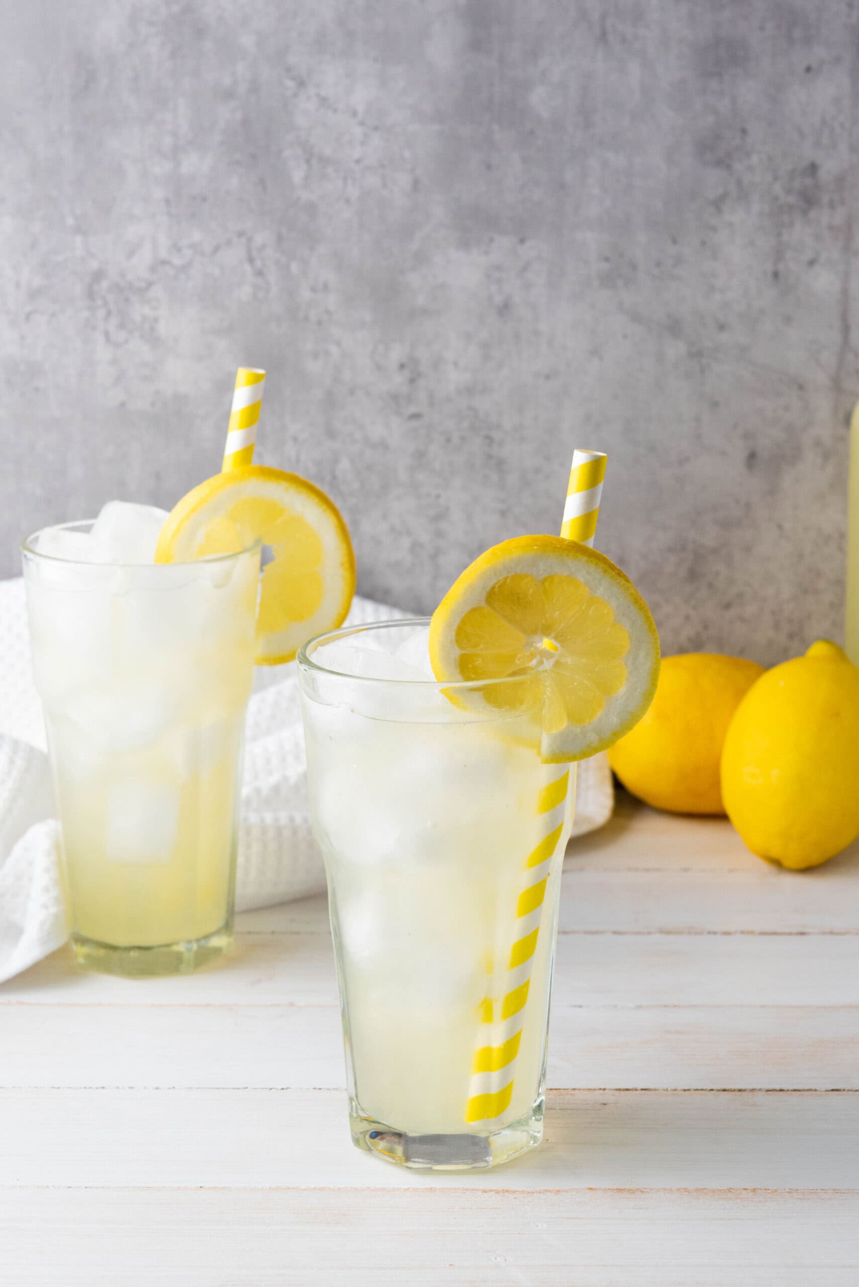 Two glasses of iced lemonade with lemon slices and yellow striped straws are pictured, alongside whole lemons in the background&mdash;perfect for enjoying a refreshing homemade lemonade recipe.