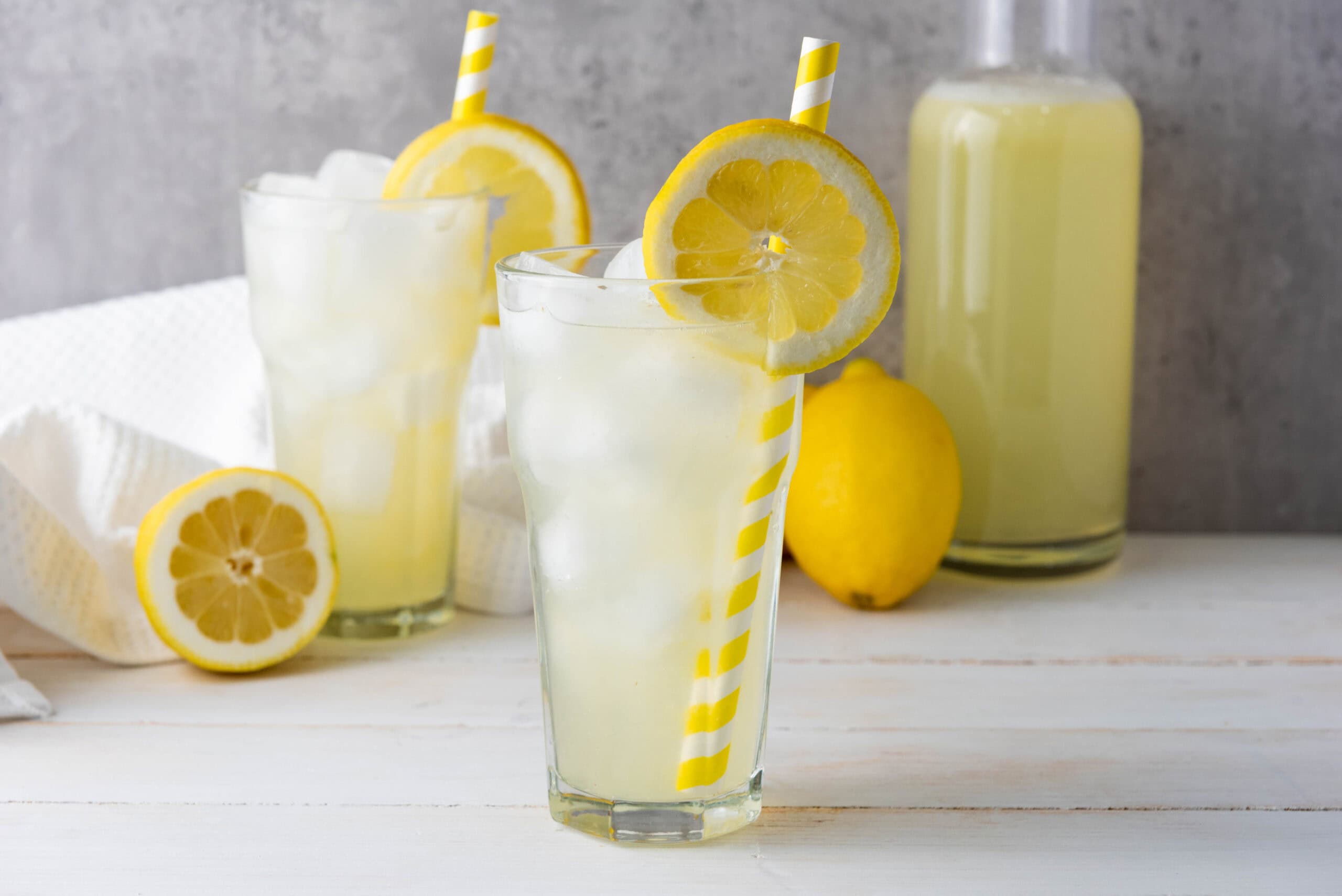 Two glasses of iced homemade lemonade with lemon slices and yellow striped straws on a white table, with fresh lemons in the background.