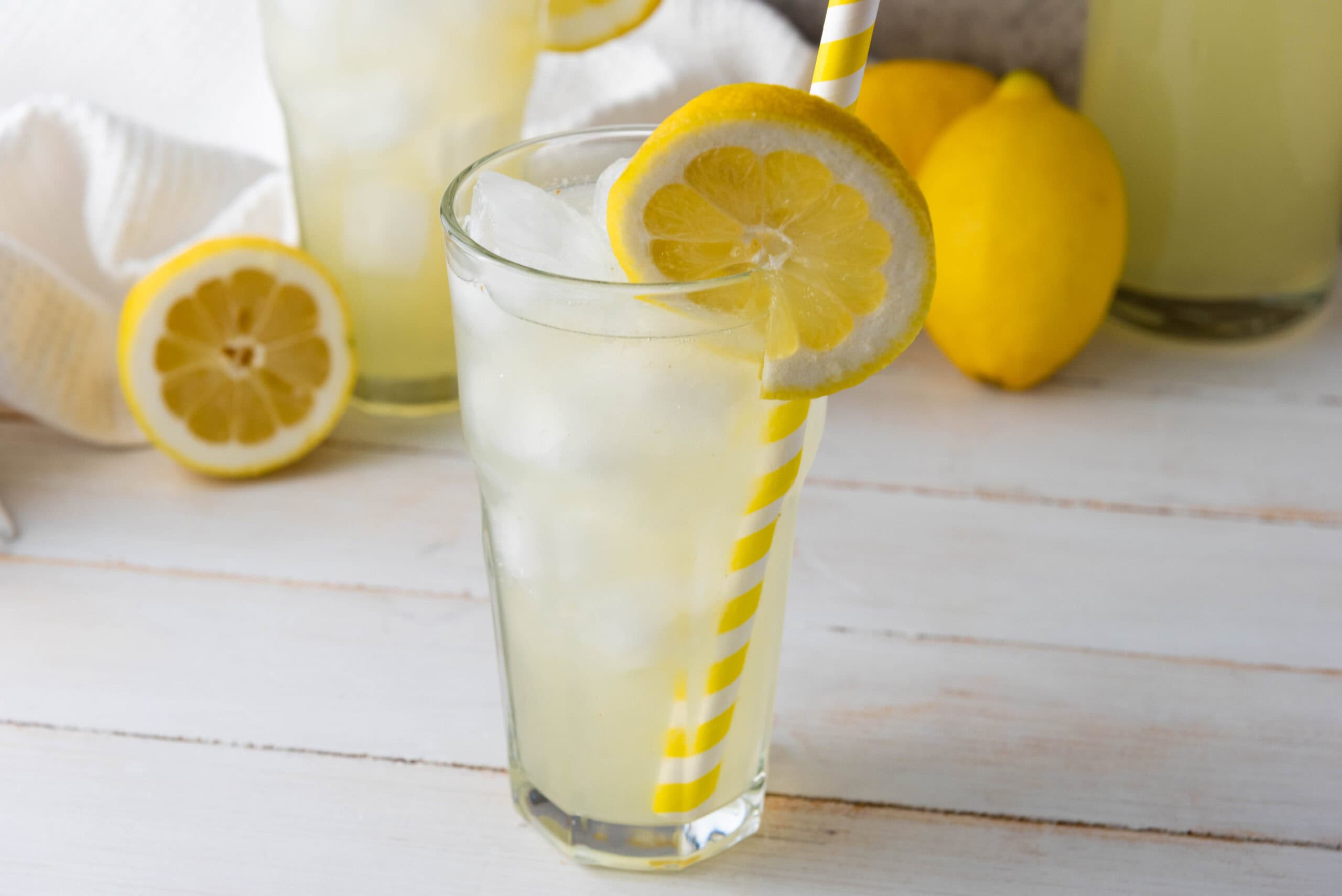 A glass of iced homemade lemonade with a lemon slice and yellow striped straw sits on a white table, with fresh lemons in the background&mdash;perfect for trying your favorite homemade lemonade recipe.