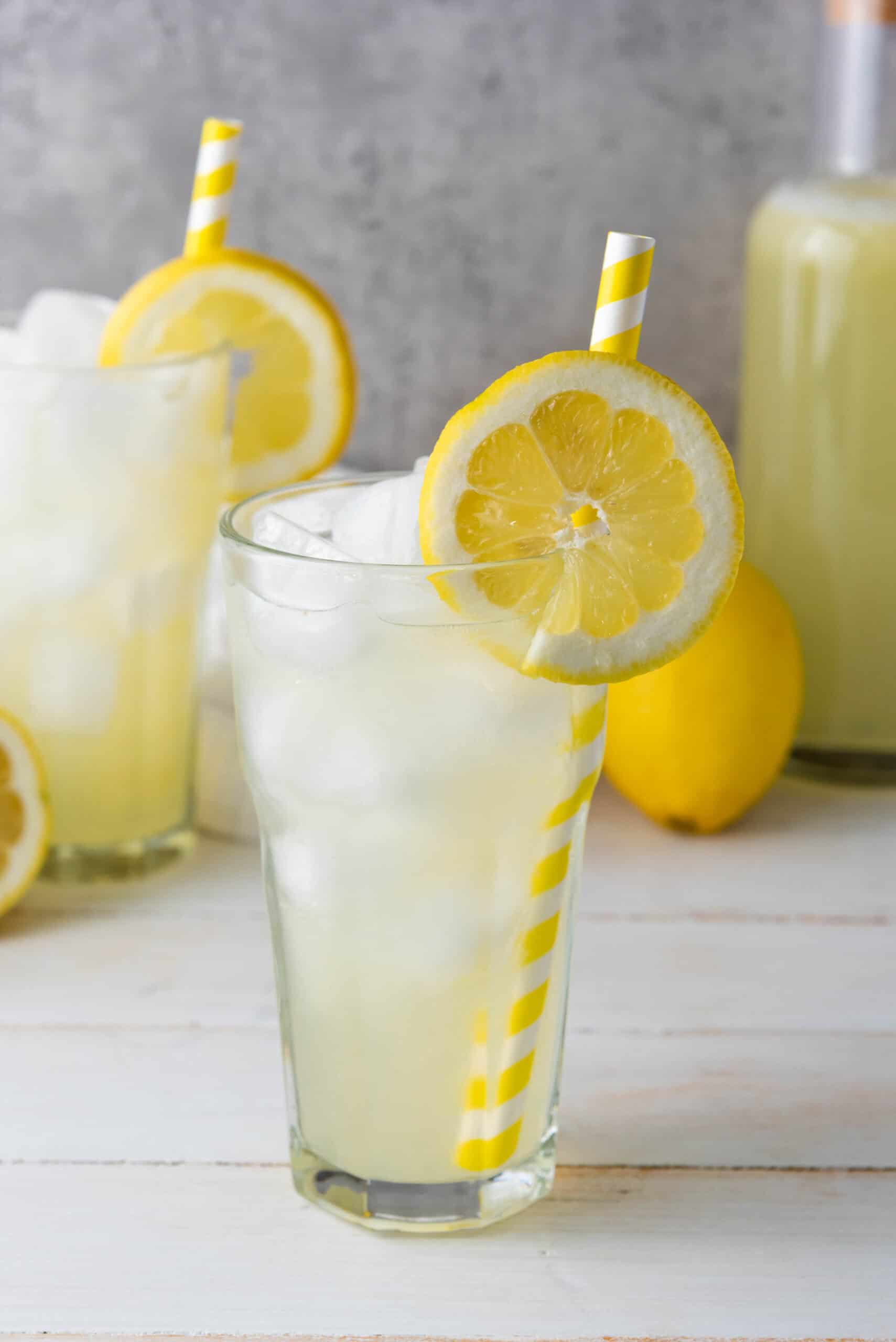 A glass of homemade lemonade with ice, a lemon slice, and a yellow striped straw sits on a white table&mdash;refreshingly simple, just like your favorite homemade lemonade recipe.