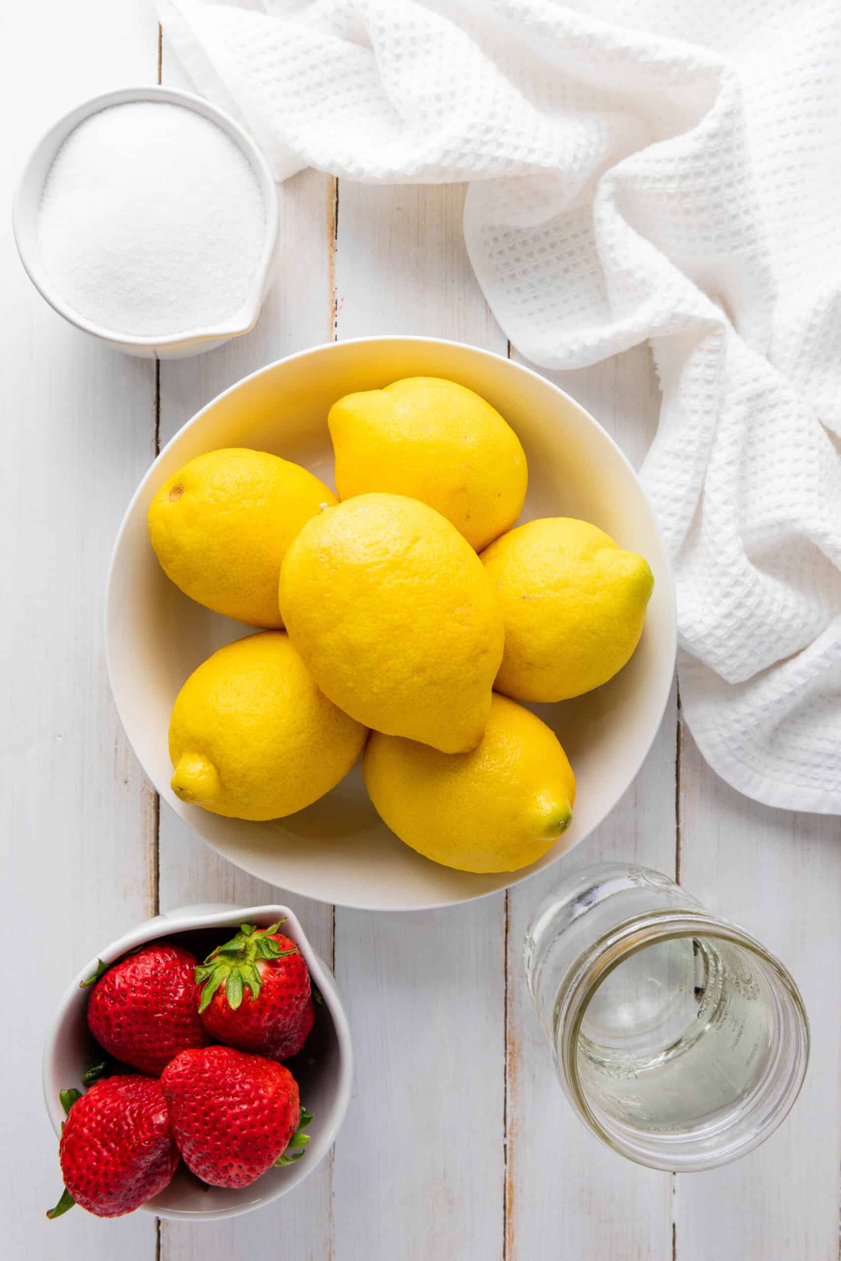 Bowl of lemons, strawberries, a jar of water, and a bowl of sugar on a white wooden table with a towel&mdash;perfect essentials for homemade strawberry lemonade.