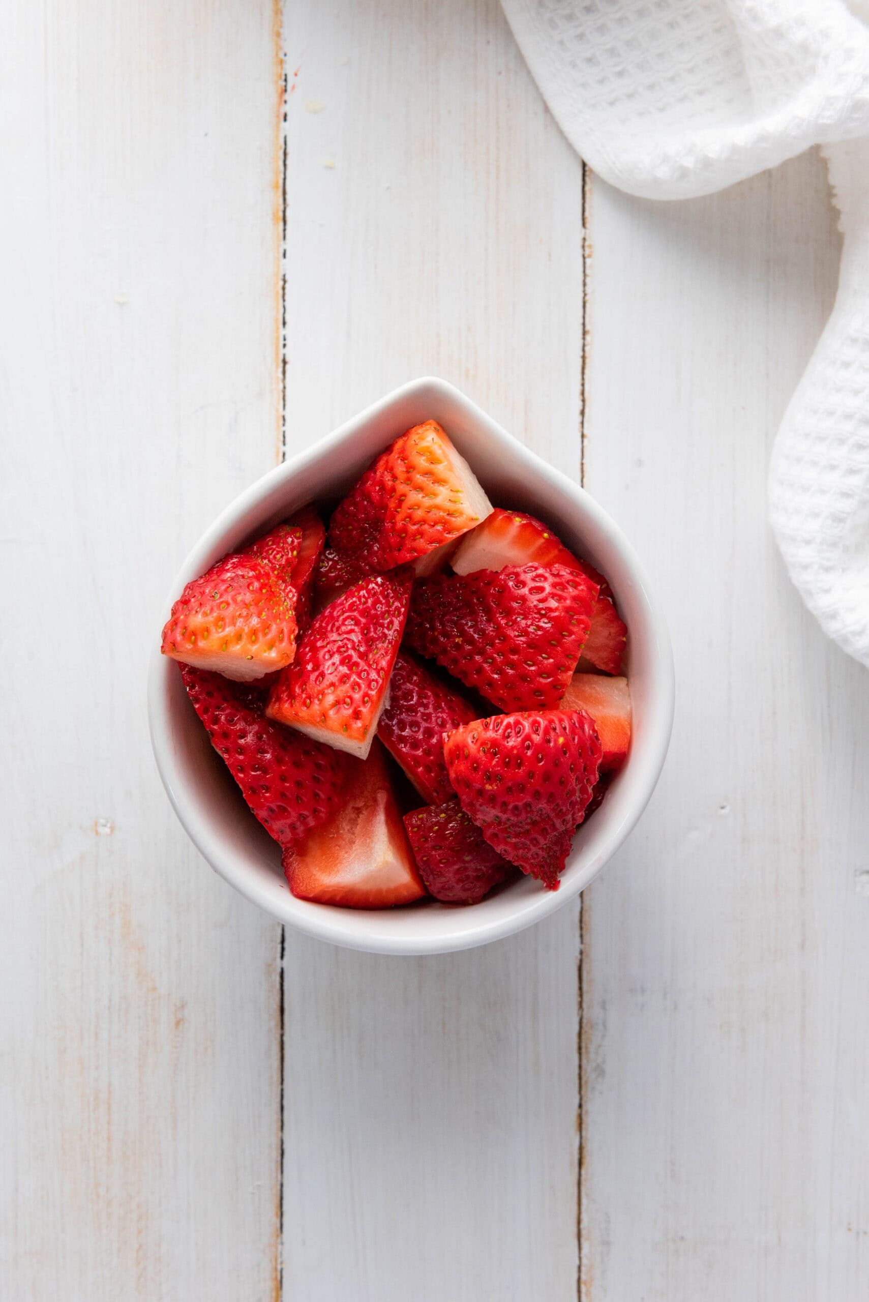 White bowl with sliced strawberries on a white wooden surface, perfect for a homemade recipe, sits next to a white textured cloth.