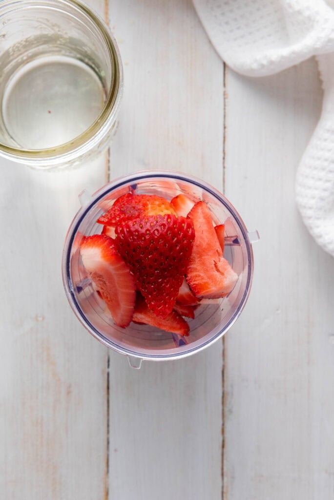 Sliced strawberries in a blender cup next to a glass jar of liquid on a white wooden surface, perfect for your Homemade Strawberry Lemonade.