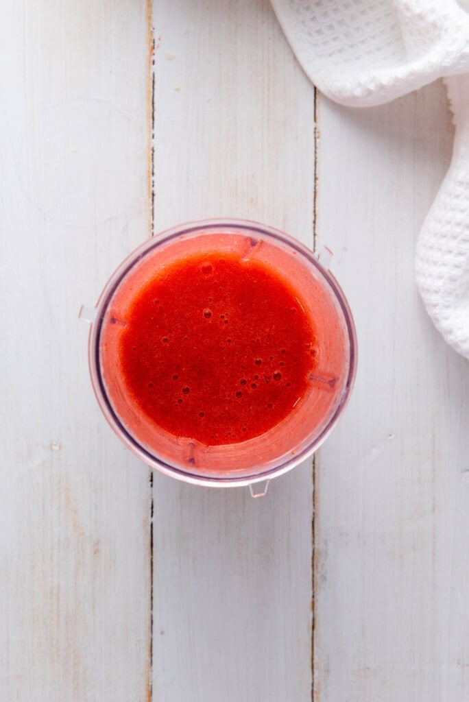 A top view of a blender cup filled with bright red smoothie on a white wooden surface, perfect for crafting a refreshing Homemade Strawberry Lemonade or trying out your favorite strawberry lemonade recipe.