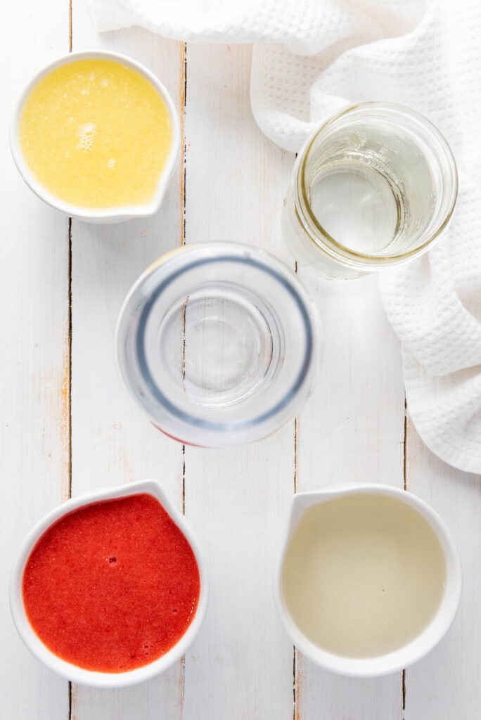 Four containers with yellow, red, and clear liquids&mdash;perfect for preparing a homemade strawberry lemonade&mdash;sit on a white wooden surface next to a white cloth.
