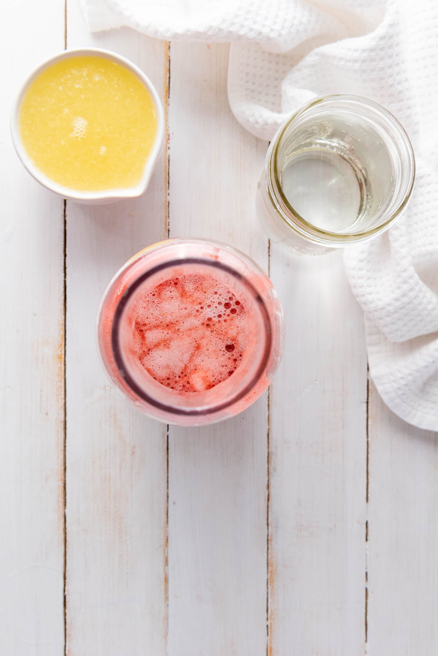 Top view of a homemade strawberry lemonade with ice, a glass of clear liquid, and a bowl of yellow liquid on a white table.