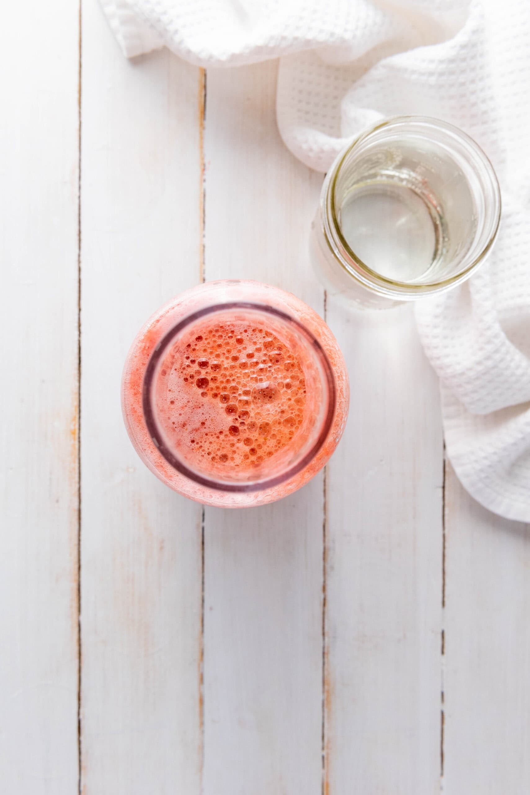 A glass of pink foamy juice, made from a Homemade Strawberry Lemonade recipe, sits beside an empty glass jar on a white wooden surface with a white cloth.