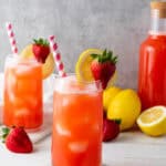 Two glasses of strawberry lemonade with ice, lemon slices, and strawberries sit beside a bottle and fresh lemons—perfect for enjoying this refreshing homemade lemonade recipe.