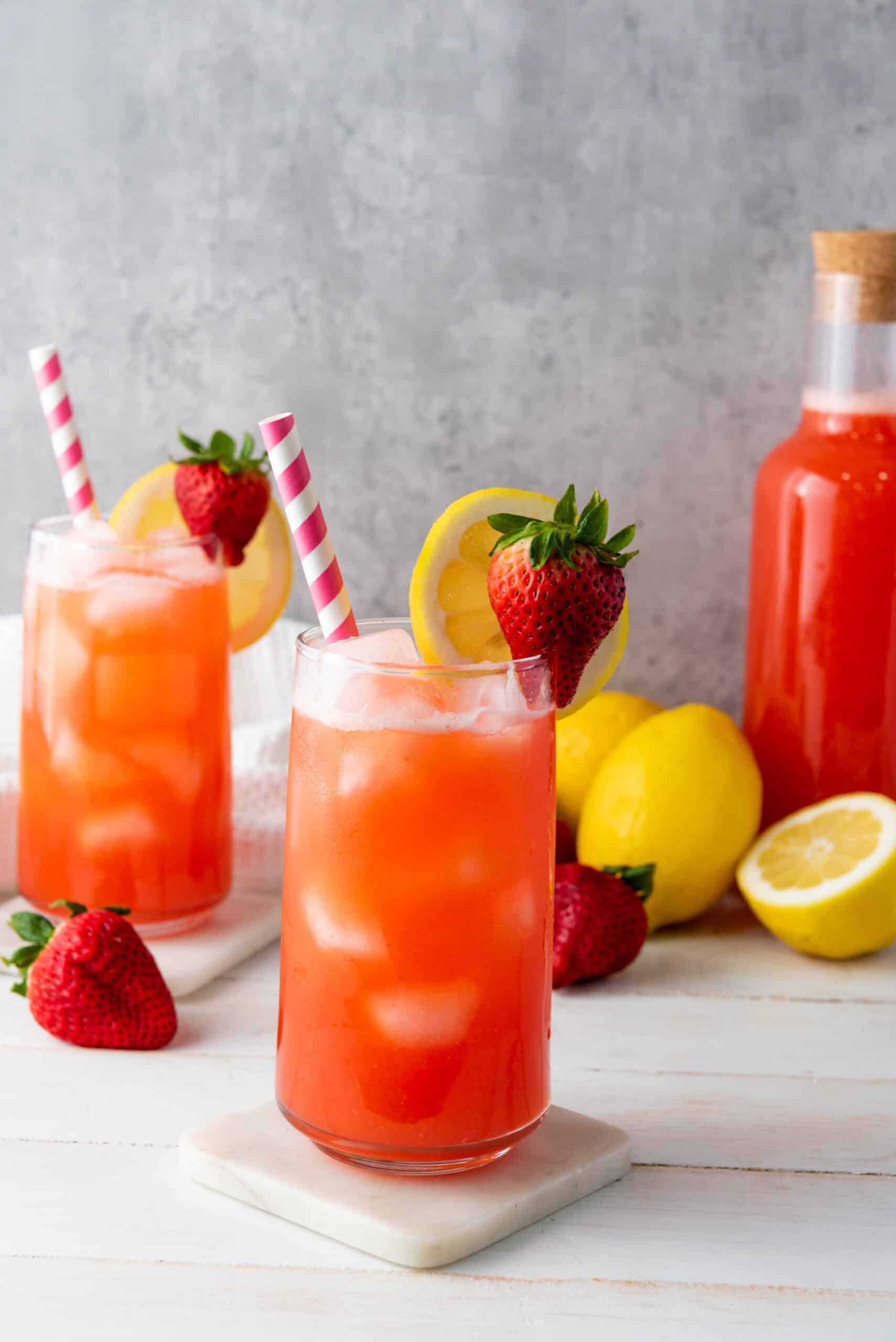Two glasses of strawberry lemonade with ice, lemon slices, and strawberries sit beside a bottle and fresh lemons&mdash;perfect for enjoying this refreshing homemade lemonade recipe.