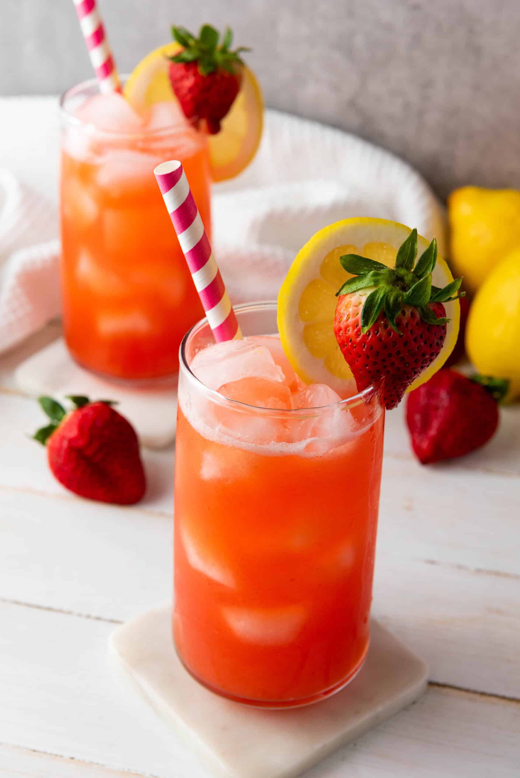 A glass of iced strawberry lemonade with a striped straw, garnished with a lemon slice and strawberry&mdash;perfect for sipping on a summer day. Try this homemade lemonade recipe for a refreshing twist!.