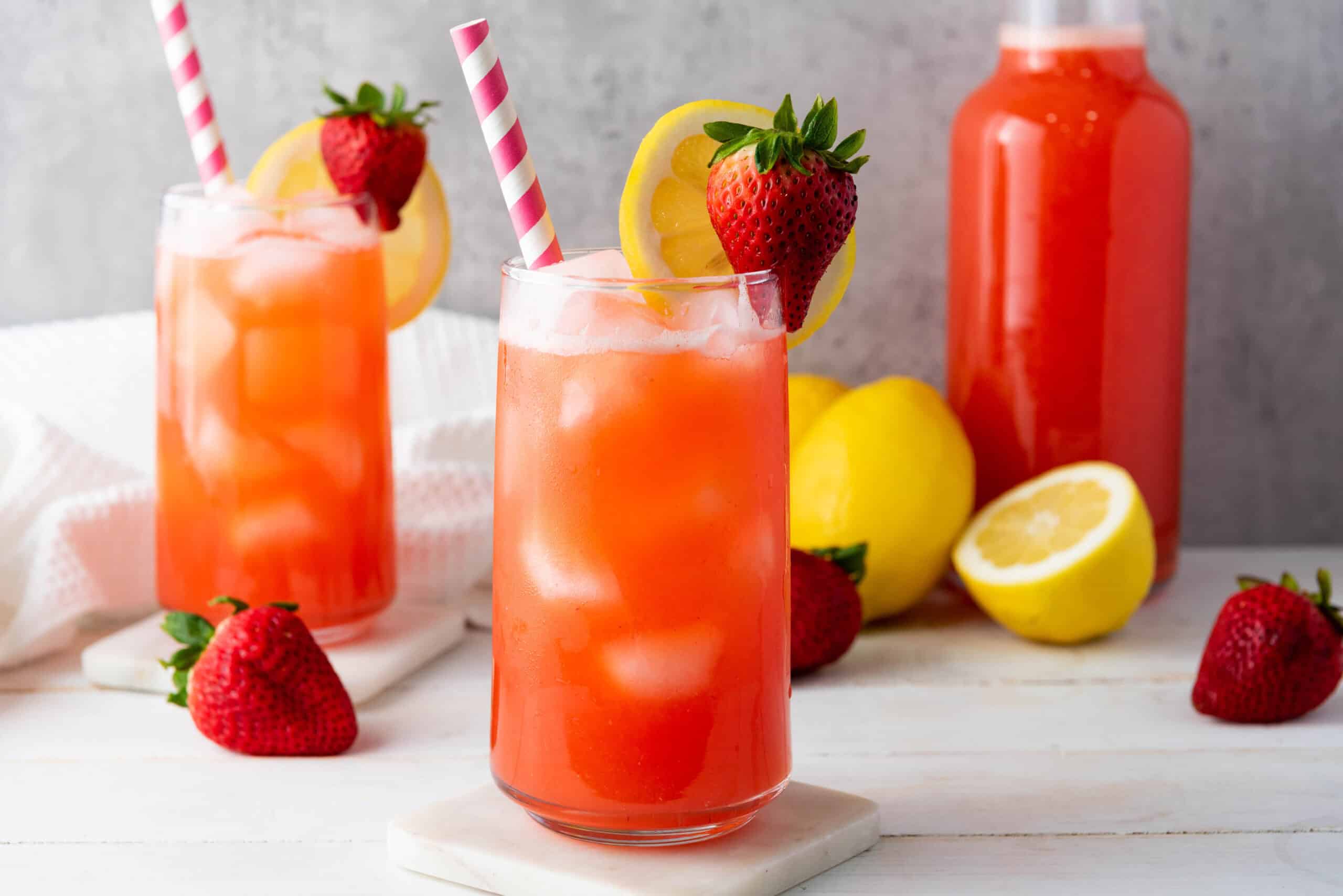 Two glasses of homemade strawberry lemonade with ice, lemon slices, and strawberries sit alongside a bottle and fresh fruit in the background. Perfect for trying your own recipe!.