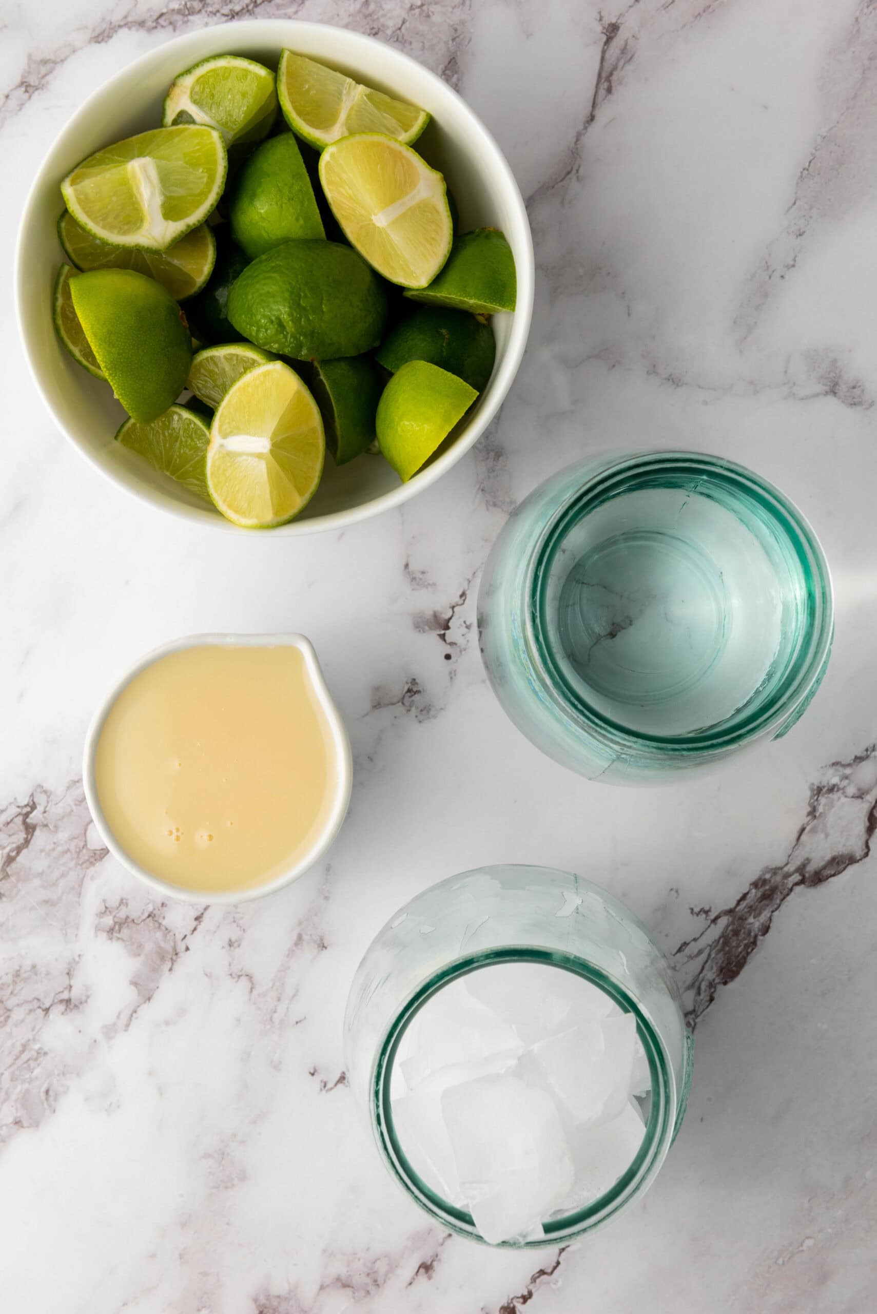 Bowl of cut limes, a cup of condensed milk, a glass of water, and a glass of ice on a marble surface&mdash;ingredients for Brazilian Limeade, a refreshing Brazilian beverage.