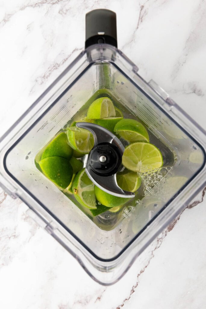 Top view of quartered limes and water in a blender on a white marble surface, perfect for preparing a refreshing Brazilian Limeade recipe.