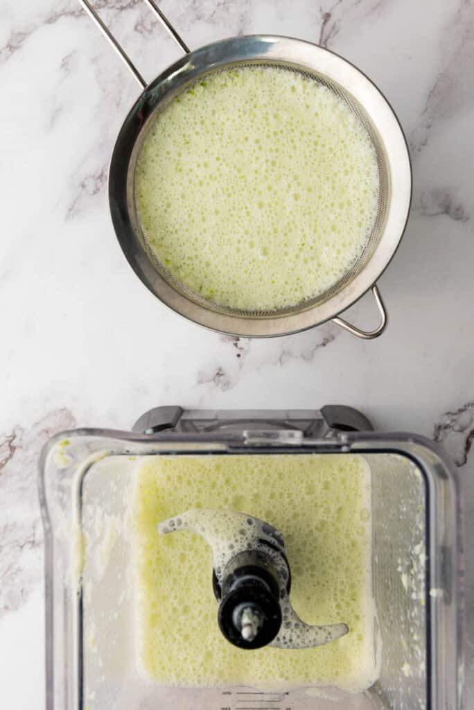 A metal strainer with green liquid, likely Brazilian Limeade, above a blender containing the same foamy green mixture on a marble counter.