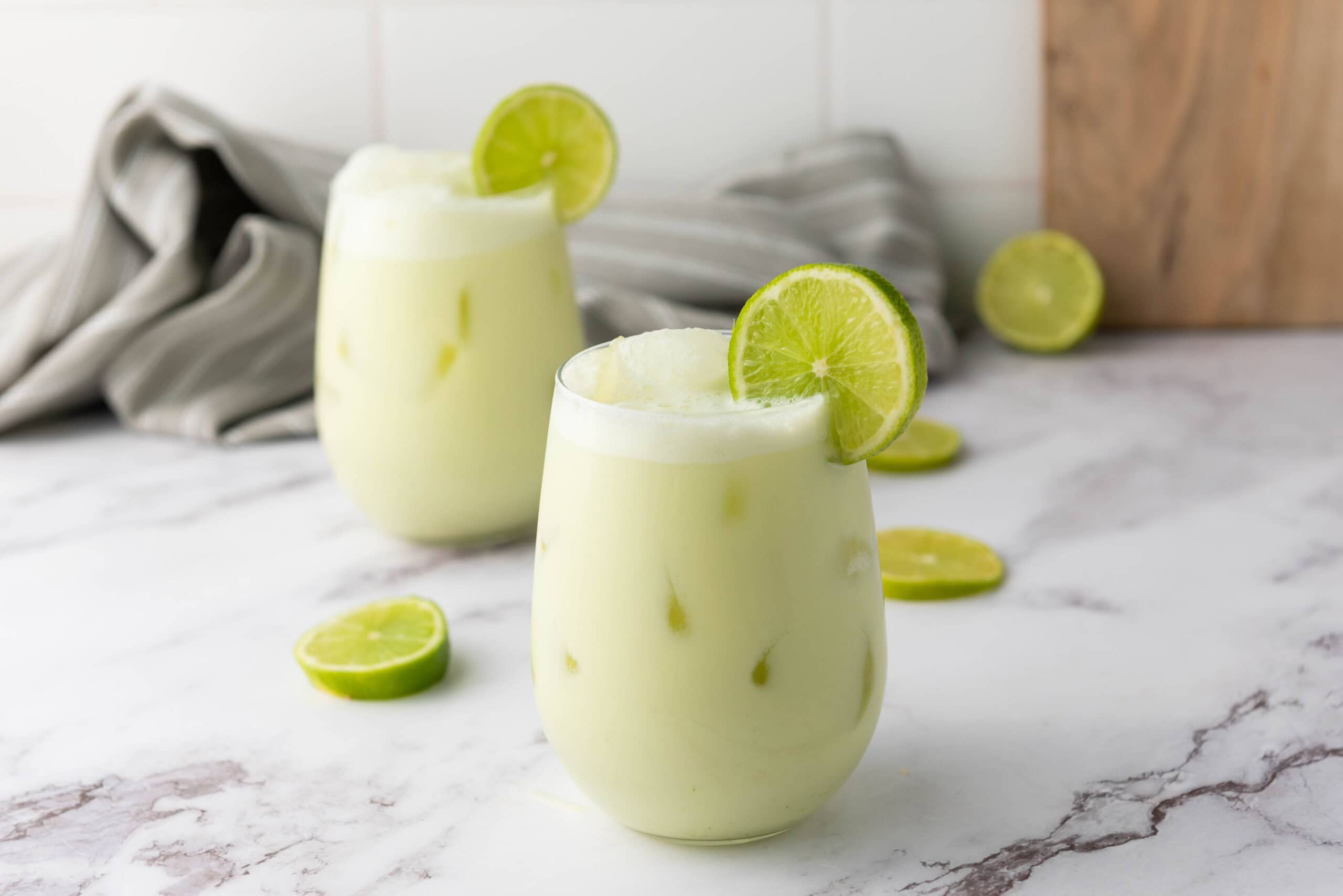 Two glasses of iced creamy Brazilian Limeade, each garnished with a lime slice, sit on a marble counter&mdash;a refreshing Brazilian drink perfect for summer.