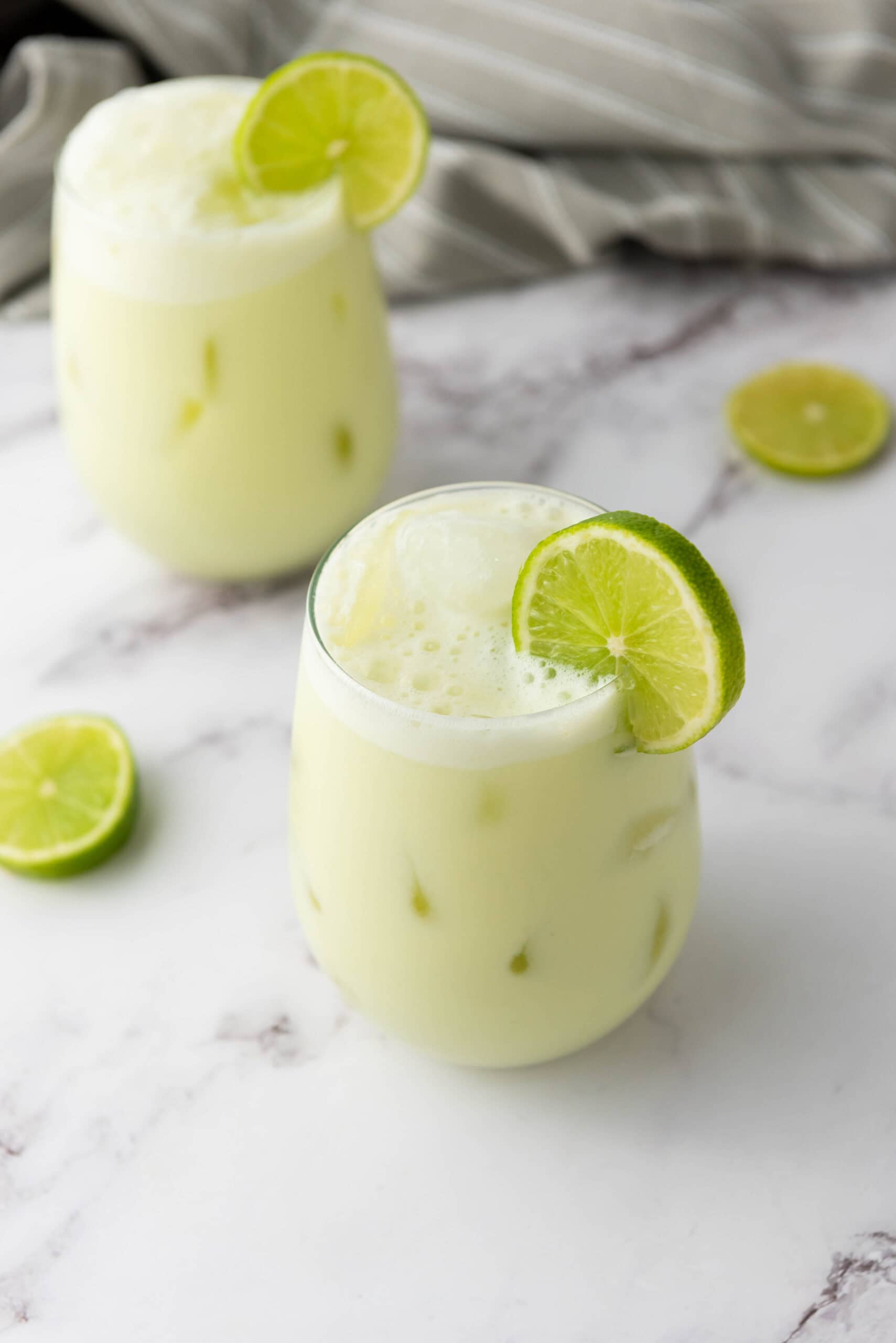 Two glasses of iced, frothy Brazilian Limeade&mdash;a classic Brazilian beverage&mdash;garnished with lime slices sit on a marble surface, surrounded by fresh limes.
