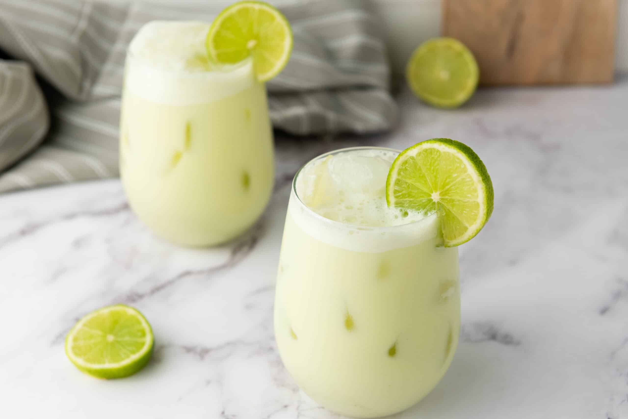 Two glasses of refreshing Brazilian Limeade are garnished with lime slices on a marble surface, with extra lime rounds nearby. This vibrant Brazilian beverage is perfect for cooling off on a hot day.