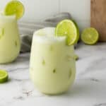 A glass of iced creamy green Brazilian Limeade, garnished with a lime slice, sits invitingly on a marble counter.