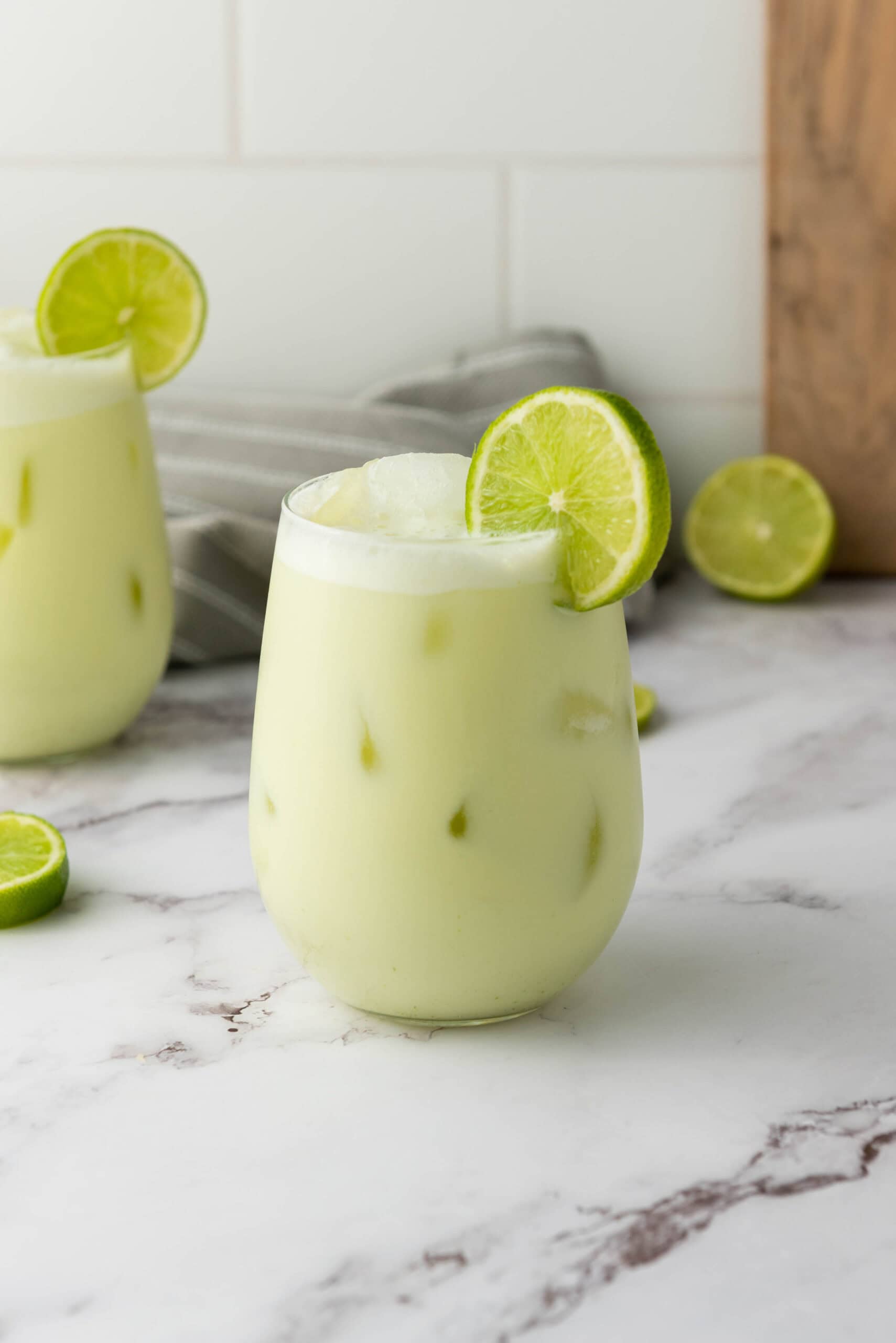 A glass of iced creamy green Brazilian Limeade, garnished with a lime slice, sits invitingly on a marble counter.