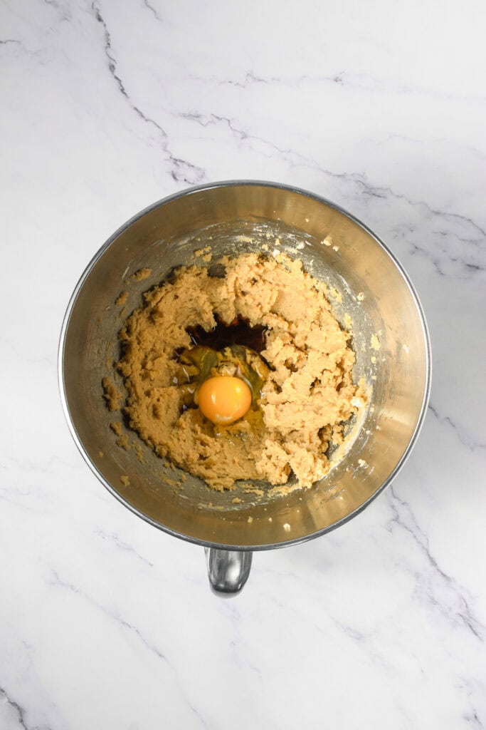 A mixing bowl with Crumbl copycat cookie dough, an egg, and vanilla extract on a marble countertop.