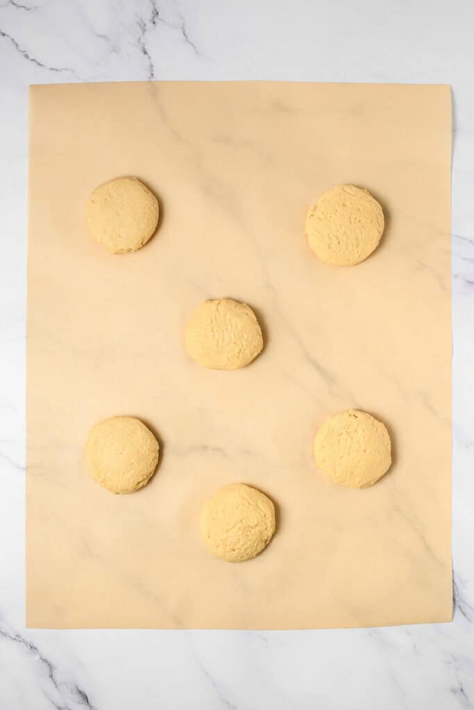 Six unbaked cookie dough cookies are arranged on parchment paper atop a marble surface, perfect for trying out your favorite cookie recipe or making a Crumbl copycat treat at home.