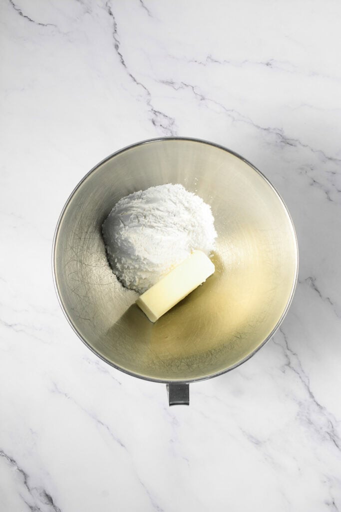 A stick of butter and powdered sugar in a metal mixing bowl on a white marble surface, ready to be whipped into Crumbl copycat cookie dough cookies.