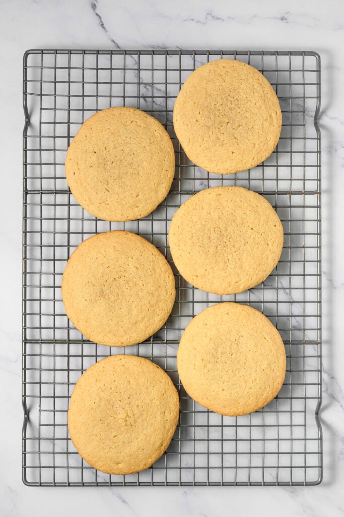 Six round cookie dough cookies cooling on a wire rack over a white marble surface, perfect for anyone craving a Crumbl copycat treat.