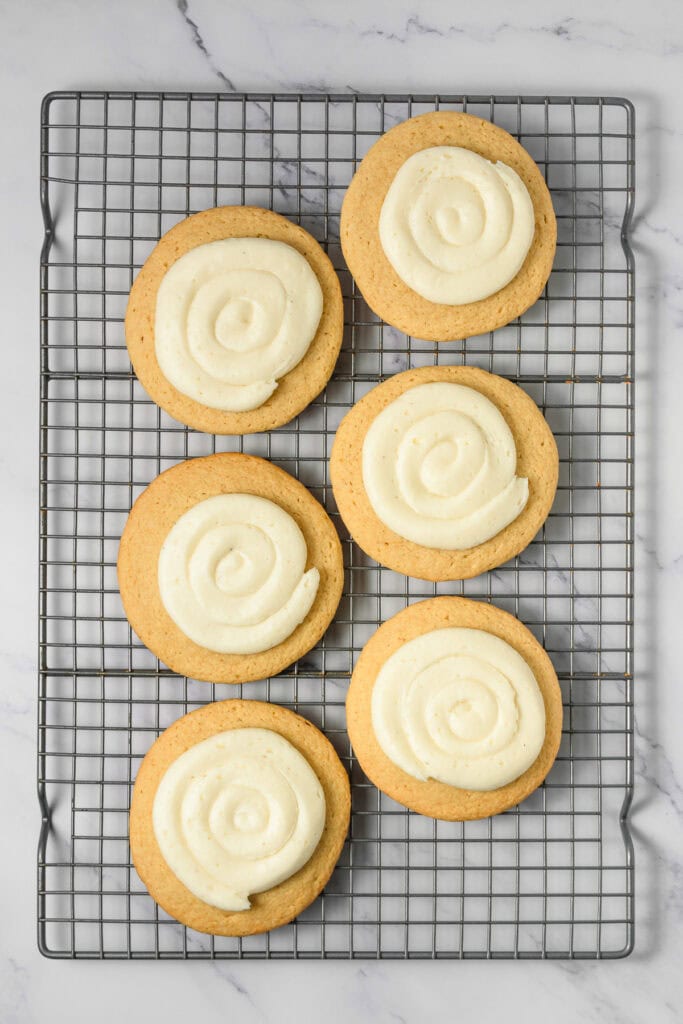 Six frosted sugar cookies with swirled icing, inspired by a Crumbl copycat cookie recipe, arranged on a cooling rack over a marble surface.