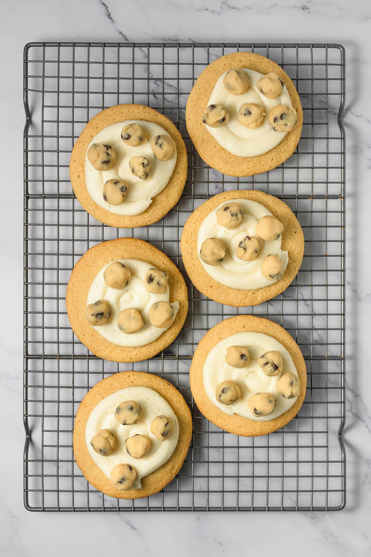 Six cookie dough cookies with white frosting and chocolate chip cookie dough balls sit on a cooling rack, top view&mdash;a perfect Crumbl copycat treat.