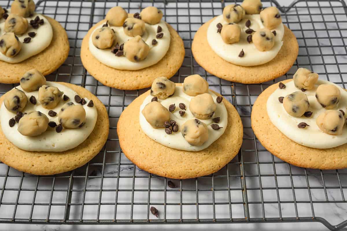 Six cookie dough cookies with frosting, topped with cookie dough balls and chocolate chips, rest on a cooling rack&mdash;just like your favorite Crumbl copycat treats.