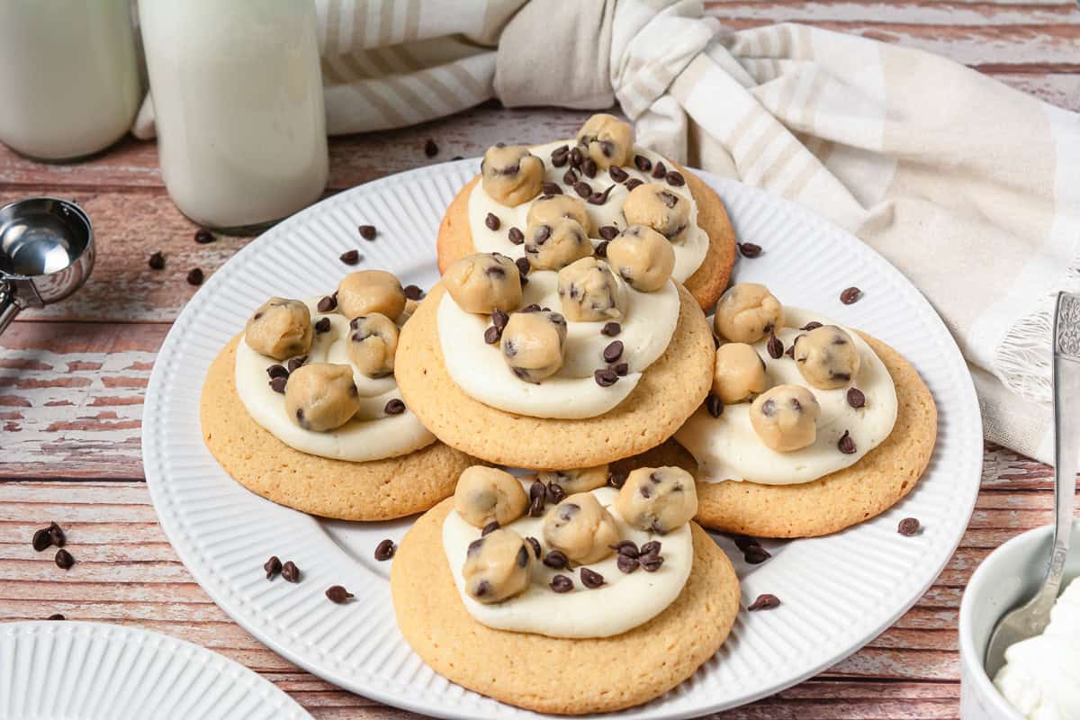 Frosted cookie dough cookies topped with chocolate chips and cookie dough bites, these Crumbl copycat treats are served on a white plate.