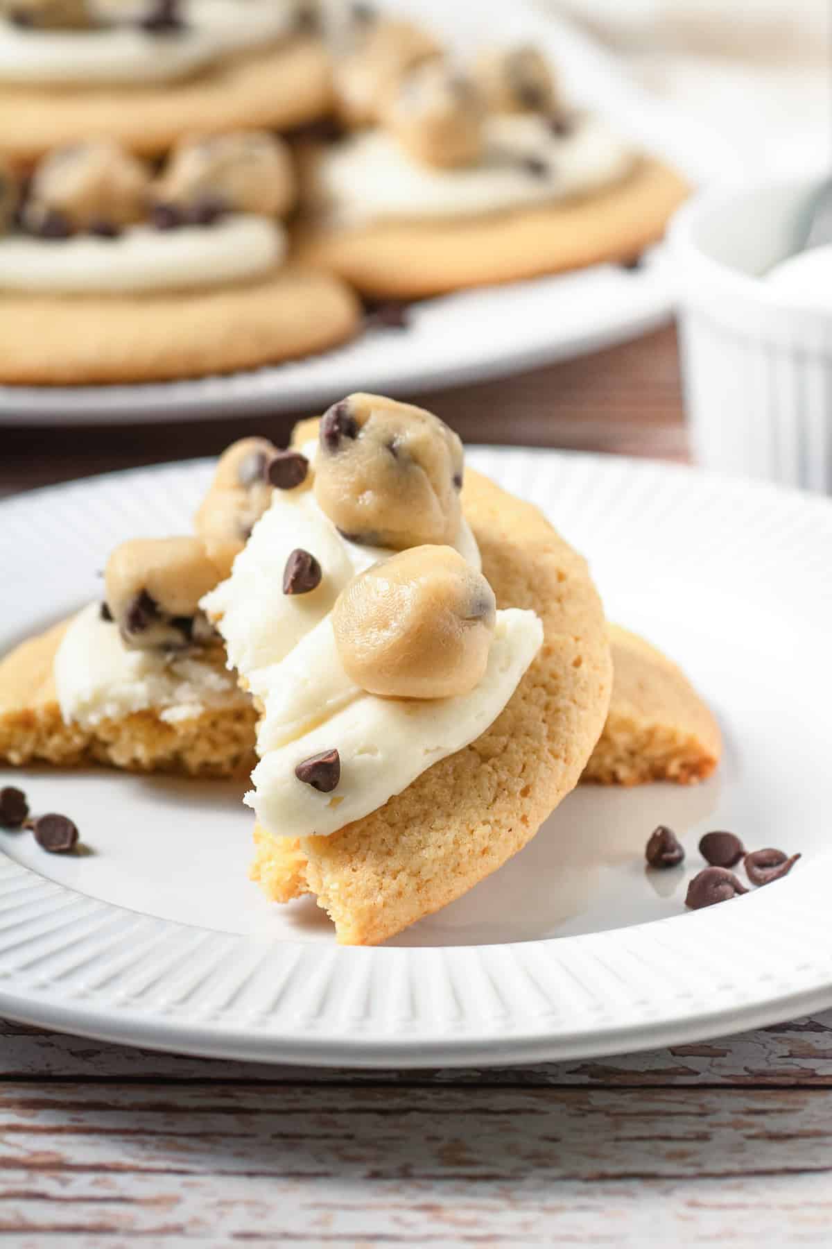 A Crumbl copycat cookie topped with frosting and cookie dough bites, garnished with chocolate chips on a white plate&mdash;the ultimate treat for cookie dough cookies lovers.