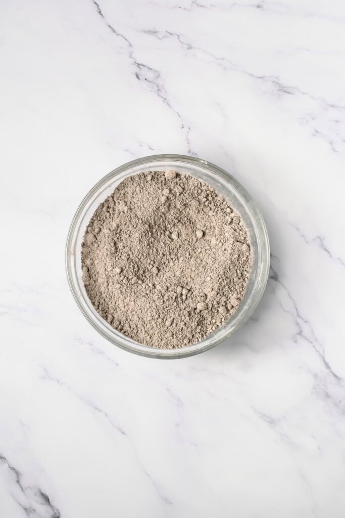 A glass bowl filled with light brown Crumbl Copycat Oreo Cookies powder sits on a white marble surface.