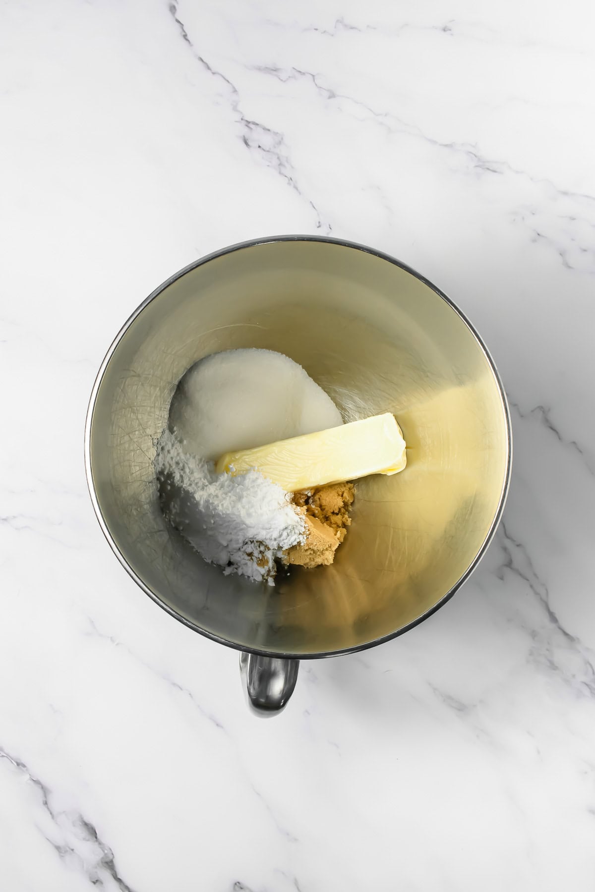 A mixing bowl with butter, white sugar, brown sugar, and powdered sugar on a marble countertop&mdash;perfect for whipping up delicious Crumbl Copycat Frosted Oreo Cookies.