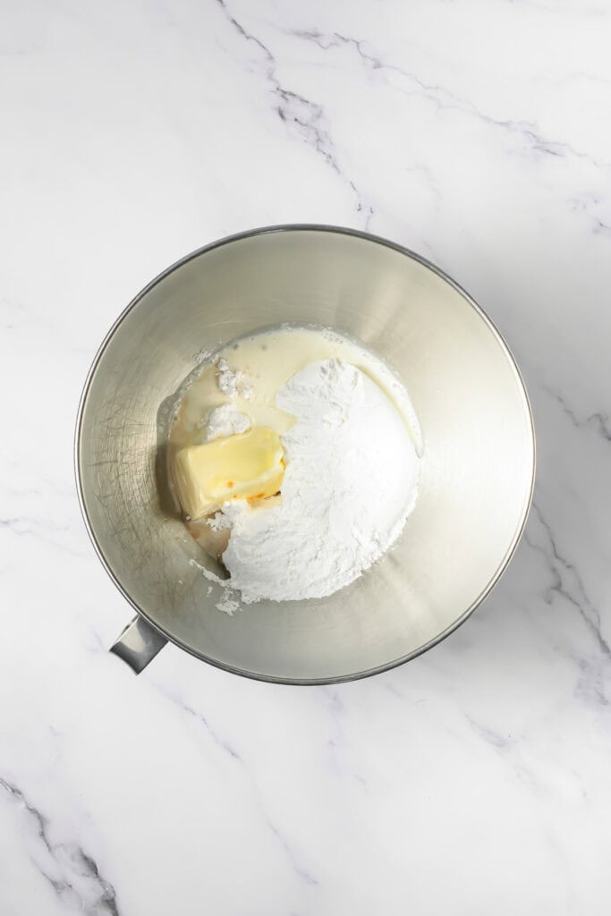 A metal mixing bowl with butter, powdered sugar, and milk sits on a white marble surface&mdash;perfect for whipping up Crumbl copycat frosted Oreo cookies.