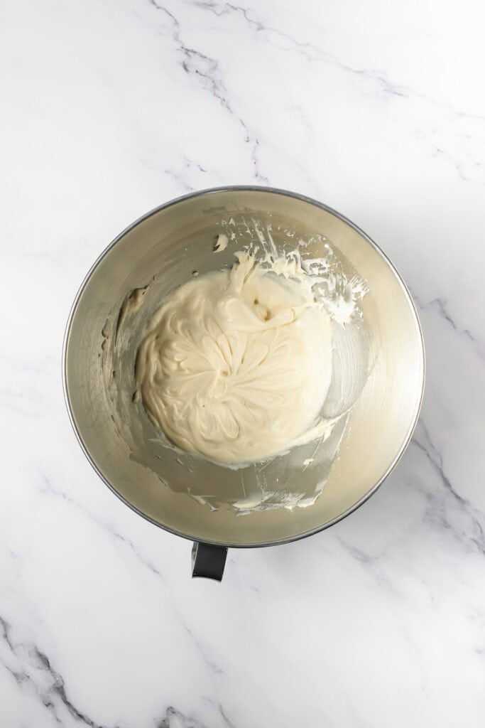 A metal mixing bowl with creamy white batter for Crumbl copycat frosted Oreo cookies sits on a white marble surface.