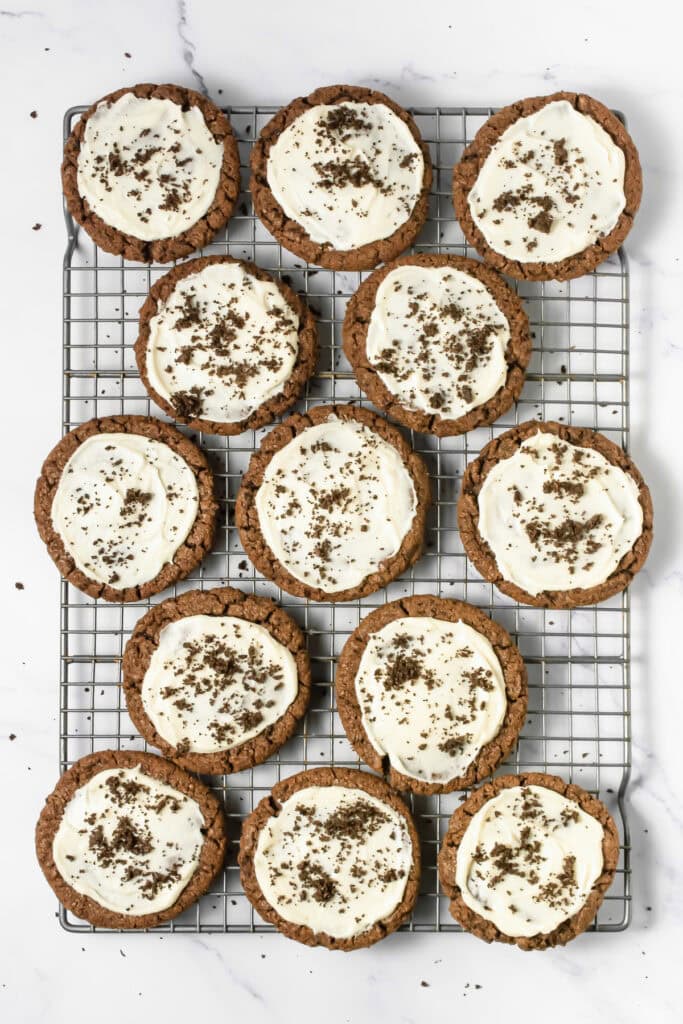 Twelve frosted Oreo cookies with white frosting and chocolate shavings rest on a cooling rack, just like a Crumbl copycat treat.