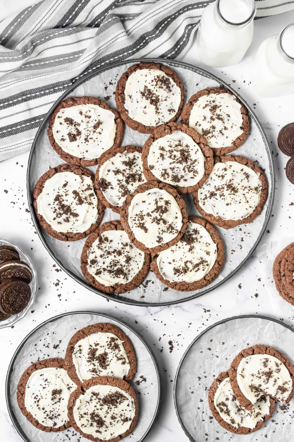 Frosted Oreo Cookies with white icing and cookie crumbs on plates, alongside milk bottles and a striped napkin, make these treats the perfect Crumbl Copycat dessert.