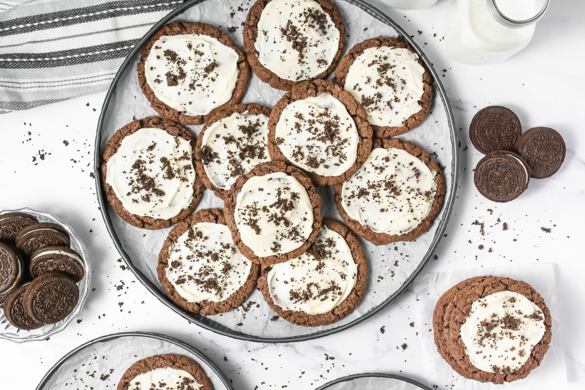 A plate of Crumbl copycat chocolate cookies with white icing and chocolate crumbs, surrounded by milk and frosted Oreo cookies.