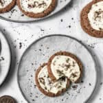 Two Crumbl Copycat frosted Oreo cookies with white frosting and cookie crumbs sit on a round plate, surrounded by more delicious cookies.