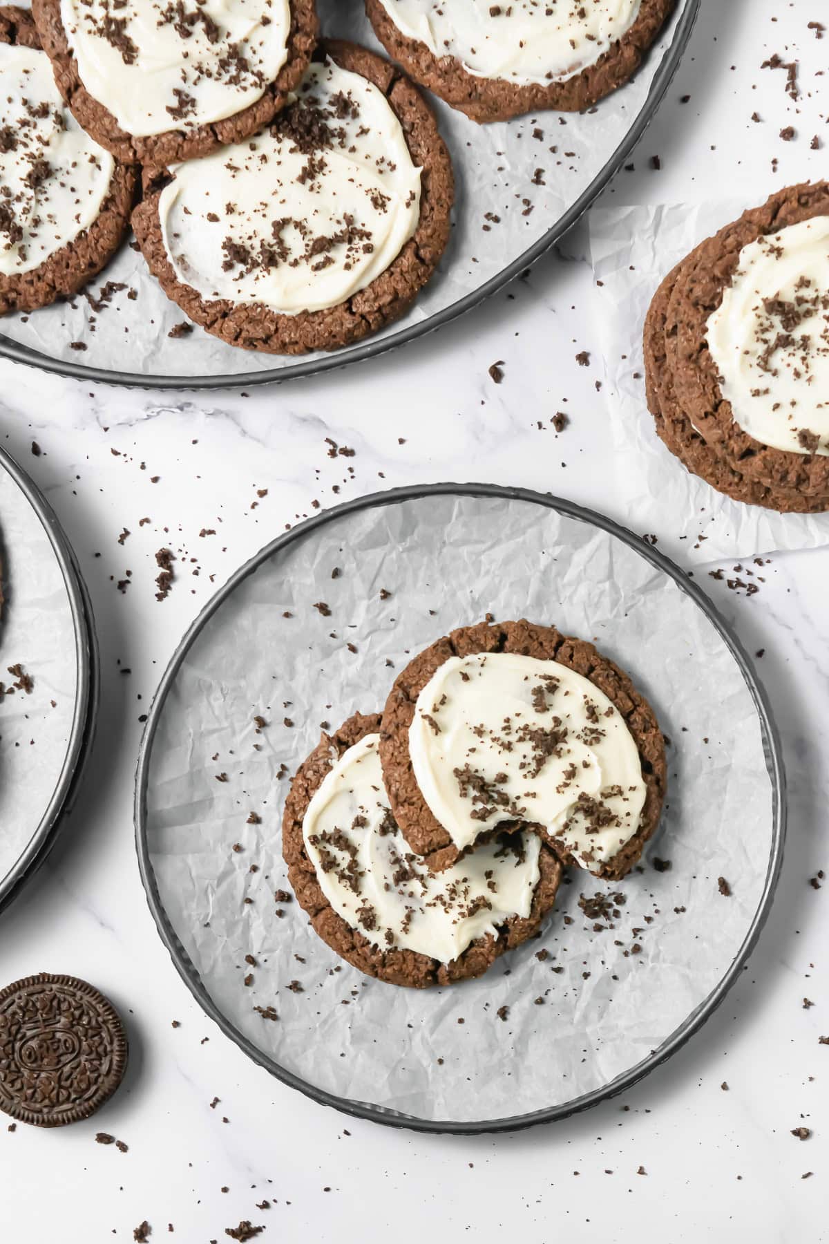 Two Crumbl Copycat frosted Oreo cookies with white frosting and cookie crumbs sit on a round plate, surrounded by more delicious cookies.