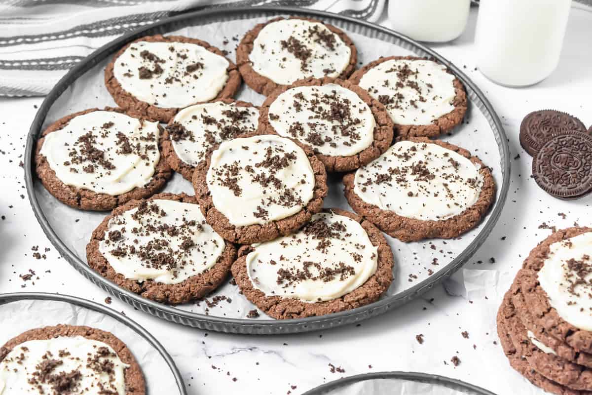 A tray of Frosted Oreo Cookies topped with white frosting and cookie crumbs, with glasses of milk nearby&mdash;your perfect Crumbl Copycat treat.