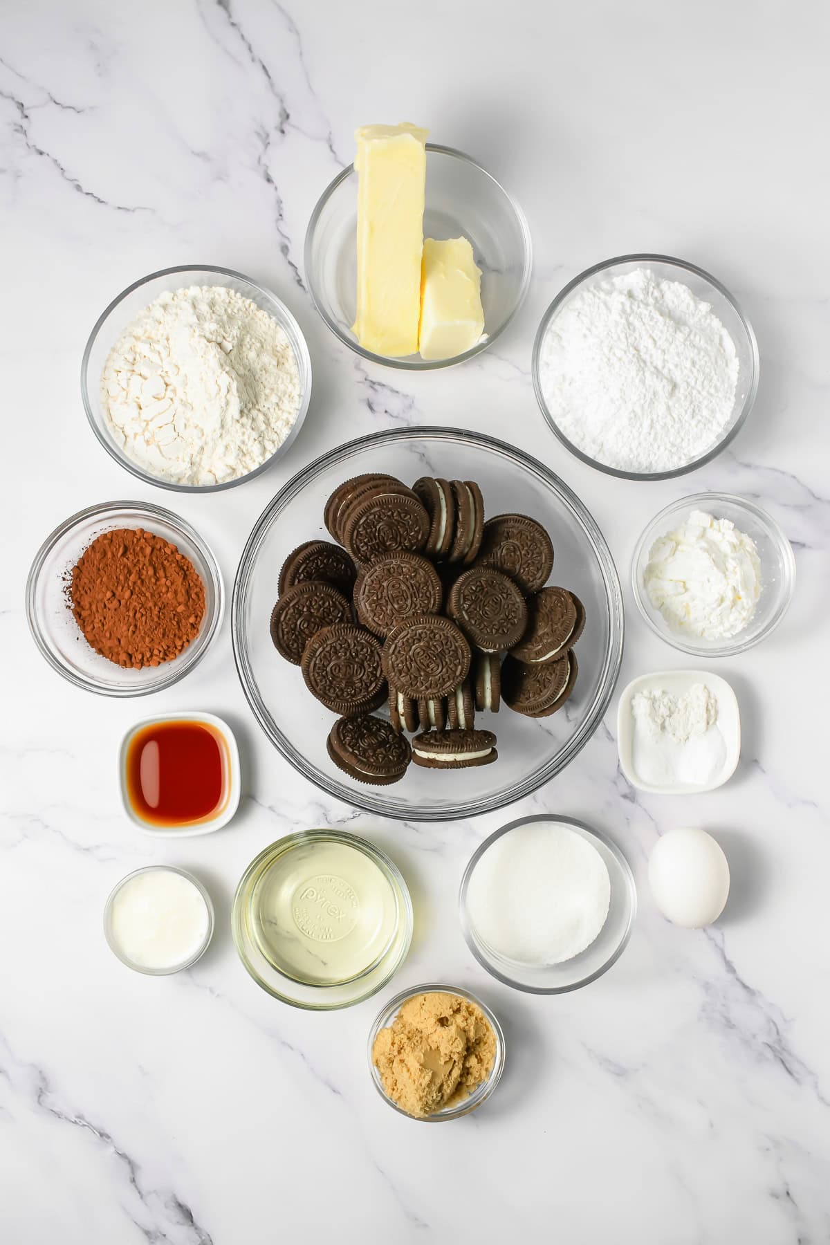 Bowls of baking ingredients, including Oreos, butter, flour, sugar, cocoa, vanilla, egg, and other powders on marble&mdash;perfect for making Crumbl Copycat Frosted Oreo Cookies.