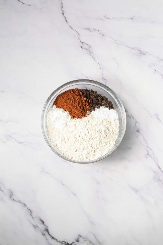 A glass bowl with flour, cocoa powder, baking soda, and salt on a white marble surface&mdash;perfect for making Crumbl copycat Frosted Oreo cookies.