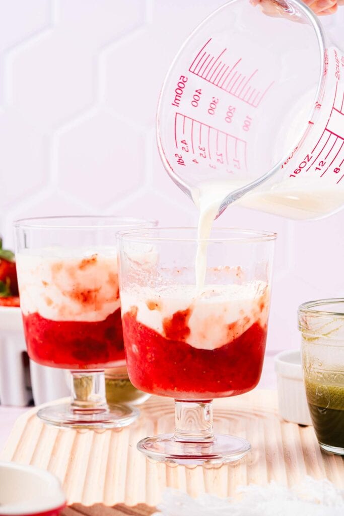 Milk is being poured into a glass with a strawberry mixture, creating the base for a delicious Strawberry Matcha Recipe; another similar glass sits nearby on the table.