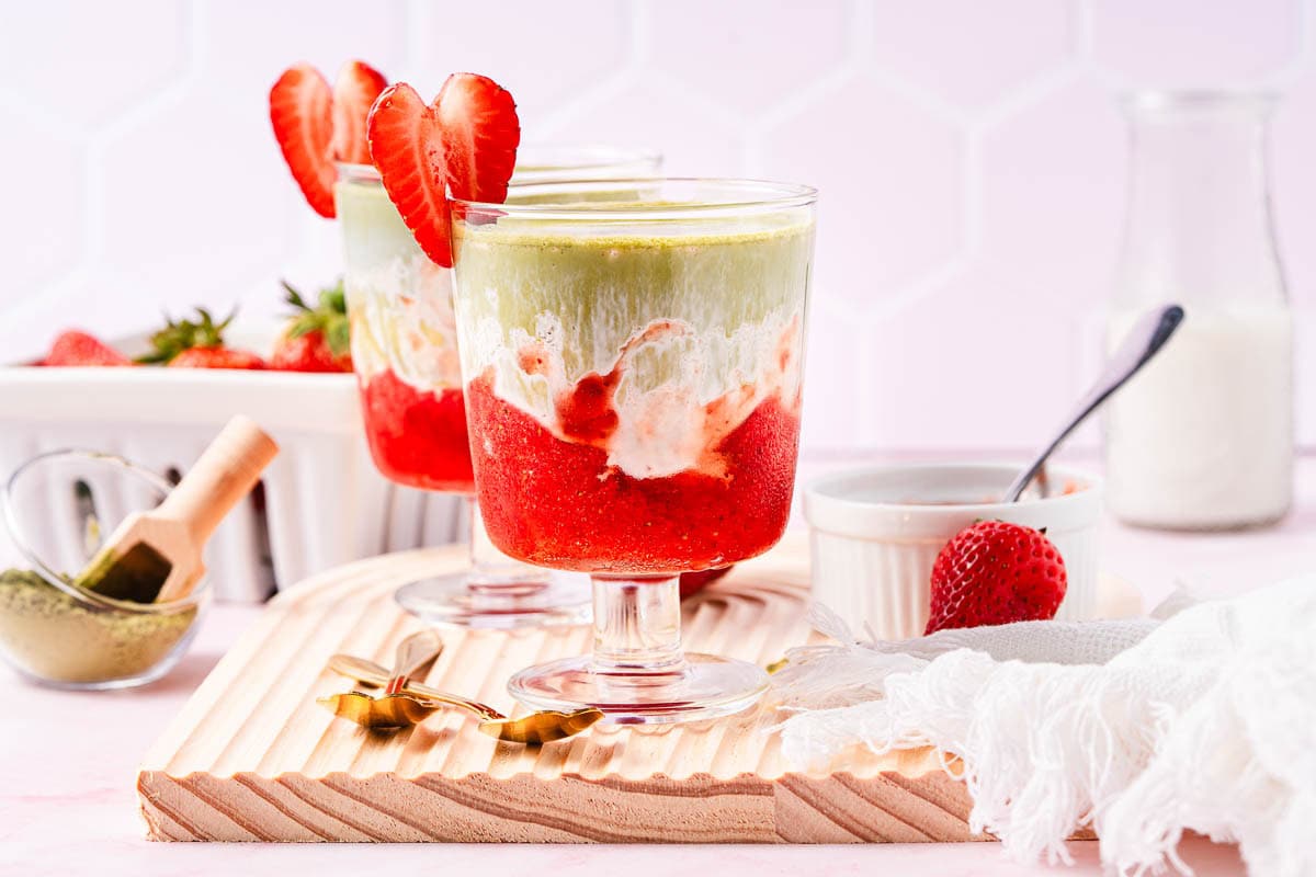Layered Strawberry Matcha drinks in glasses, garnished with fresh strawberries, on a wooden board with a white napkin&mdash;a vibrant and refreshing Strawberry Matcha Recipe perfect for any occasion.