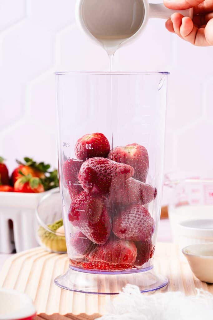 A hand pours liquid over strawberries in a blender jar on a wooden surface, preparing a delicious Strawberry Matcha recipe.
